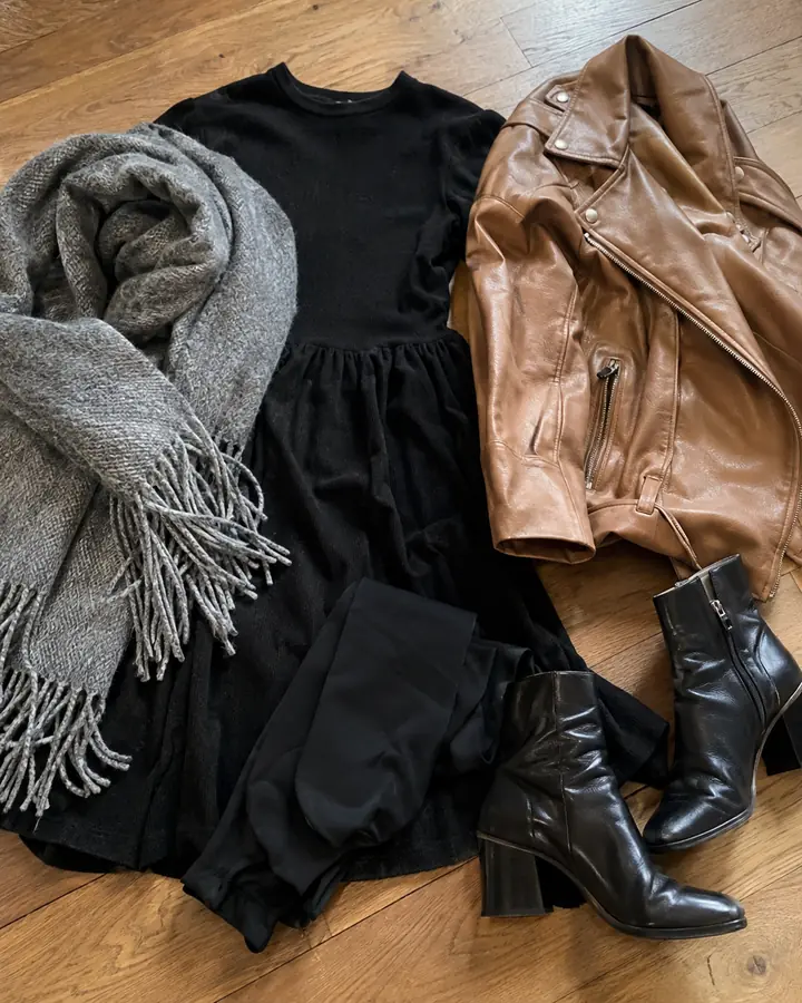 Flat lay of a black dress, black tights, ankle boots, a grey scarf and a leather jacket on a wooden floor